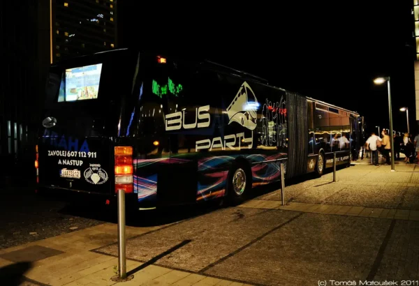 A large, dark-colored bus with "Prague XXXL Party Bus" written on the side is parked on a street at night. The bus has vibrant, colorful graphics and lights, perfect for a prague stag fun night. A group of people can be seen mingling near the door of the bus. The photo is attributed to Tomáš Malatoušek, 2011.