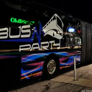 A side view of a decorated Prague XXXL Party Bus at night in Prague. The bus features colorful wave patterns, silhouettes of dancing people, and the words "BUS PARTY" prominently displayed. Bright lights on the bus reflect an energetic atmosphere perfect for a stag party. Photo credit: Tomáš Marušinec 2011.