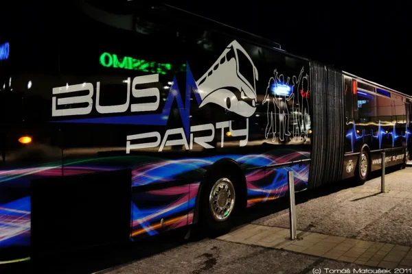 A side view of a decorated Prague XXXL Party Bus at night in Prague. The bus features colorful wave patterns, silhouettes of dancing people, and the words "BUS PARTY" prominently displayed. Bright lights on the bus reflect an energetic atmosphere perfect for a stag party. Photo credit: Tomáš Marušinec 2011.