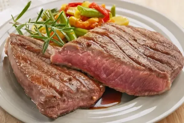 A plate with two medium-rare grilled steaks garnished with a sprig of rosemary. In the background, a colorful medley of sautéed vegetables including bell peppers and green beans completes this feast perfect for any stag party. The steaks boast visible grill marks and are slightly pink in the center.