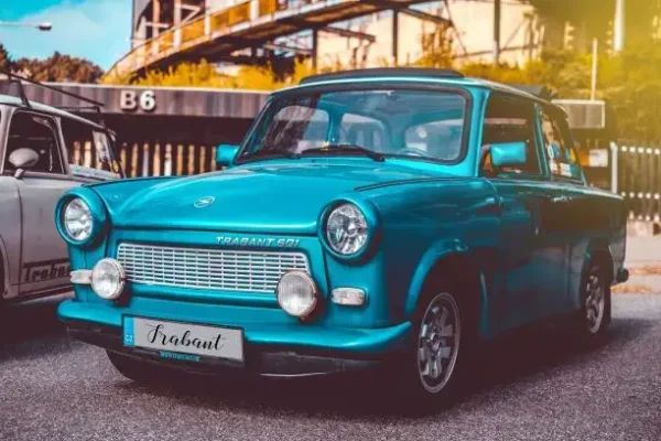 A vintage turquoise Trabant 601 car is parked on the street, a perfect backdrop for a Crazy Trabant Ride with Beer in Prague. Its classic boxy design is complemented by round headlights and a Trabant logo on the grille. A hint of another car and industrial structures in the background are visible. The license plate reads "Trabant.