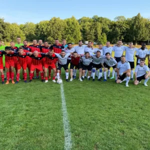 Two soccer teams pose together, smiling on a grassy field in Prague. The team on the left wears red and black uniforms, and the team on the right wears white and black uniforms. Trees and a clear blue sky are visible in the background. A Prague Football Match ball is placed at the center.