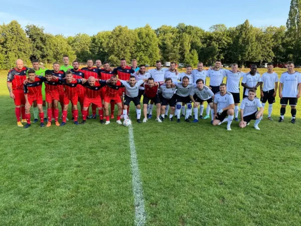 Two soccer teams pose together, smiling on a grassy field in Prague. The team on the left wears red and black uniforms, and the team on the right wears white and black uniforms. Trees and a clear blue sky are visible in the background. A Prague Football Match ball is placed at the center.