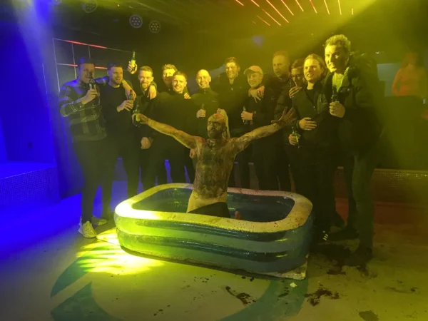 A group of men stands in a semi-circle around an inflatable kiddie pool during their Prague stag do. In the pool, a man covered in mud raises his arms triumphantly. The setting is a nightclub with colorful lighting and a vibrant atmosphere, and several of the men are holding drinks, enjoying their Prague stag activities.