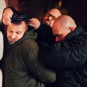 Three men in dark clothing restrain a man in a green jacket at night against a building wall. The restrained man, part of a Prank Stag Arrest & Strip, appears distressed, while the two other men seem focused, with one securing the man's hands behind his back and the other pressing him against the wall.