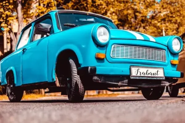 A bright blue vintage Trabant car is parked on the street with autumn foliage in the background. The image, taken from a low angle, makes the car look prominent against the golden leaves. Perfect for a Crazy Trabant Ride with Beer, this scene adds a quirky twist to your Prague stag fun. The license plate reads "Trabant.